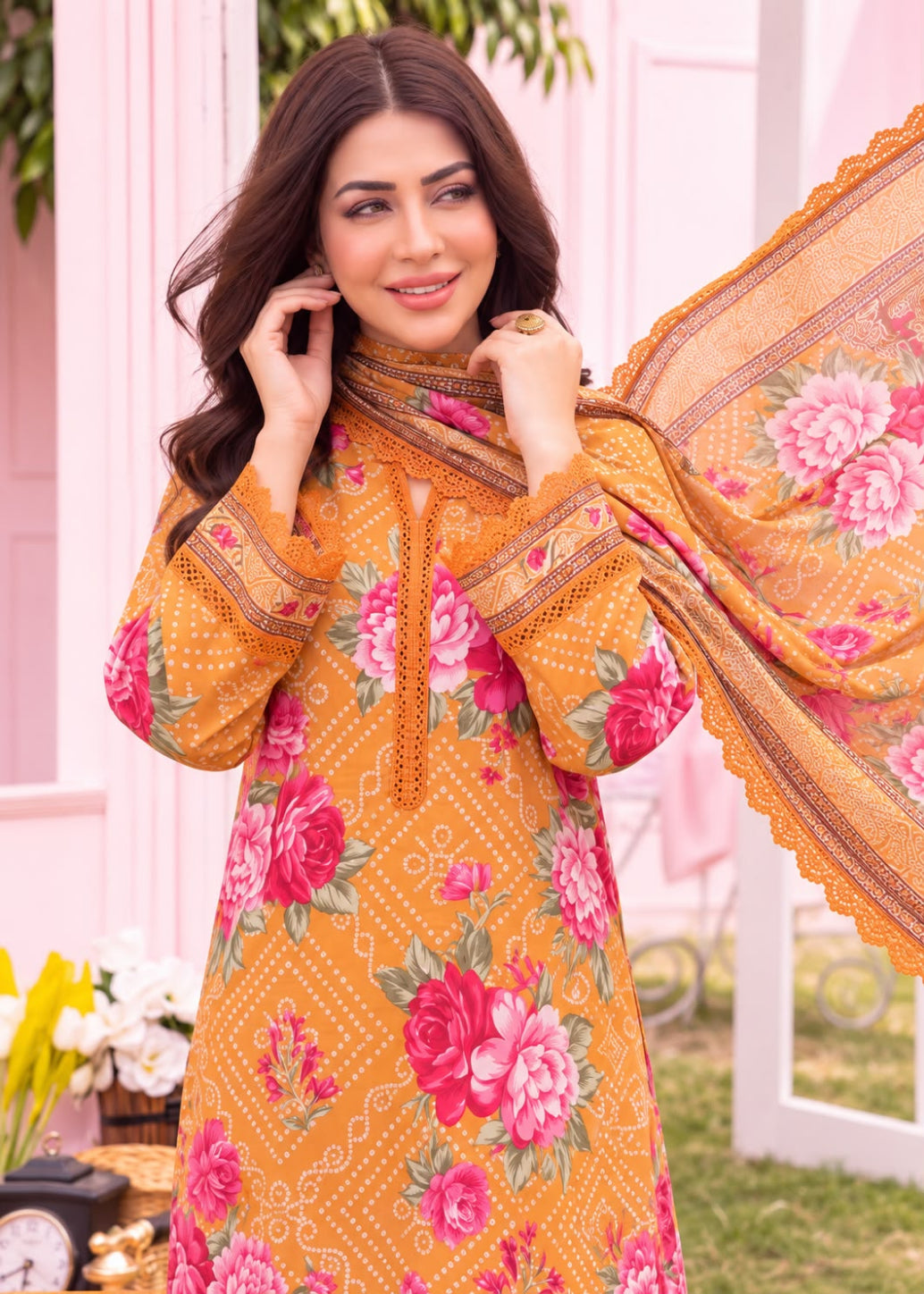 A woman models a mustard yellow 3-piece salwar kameez suit with a pink and green floral print, featuring a matching printed dupatta draped over her shoulder, set against a soft pink background.