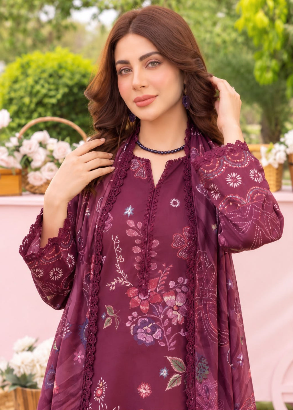 A woman wearing a deep purple embroidered kurta with a matching sheer dupatta, set against a soft pink and green garden background.