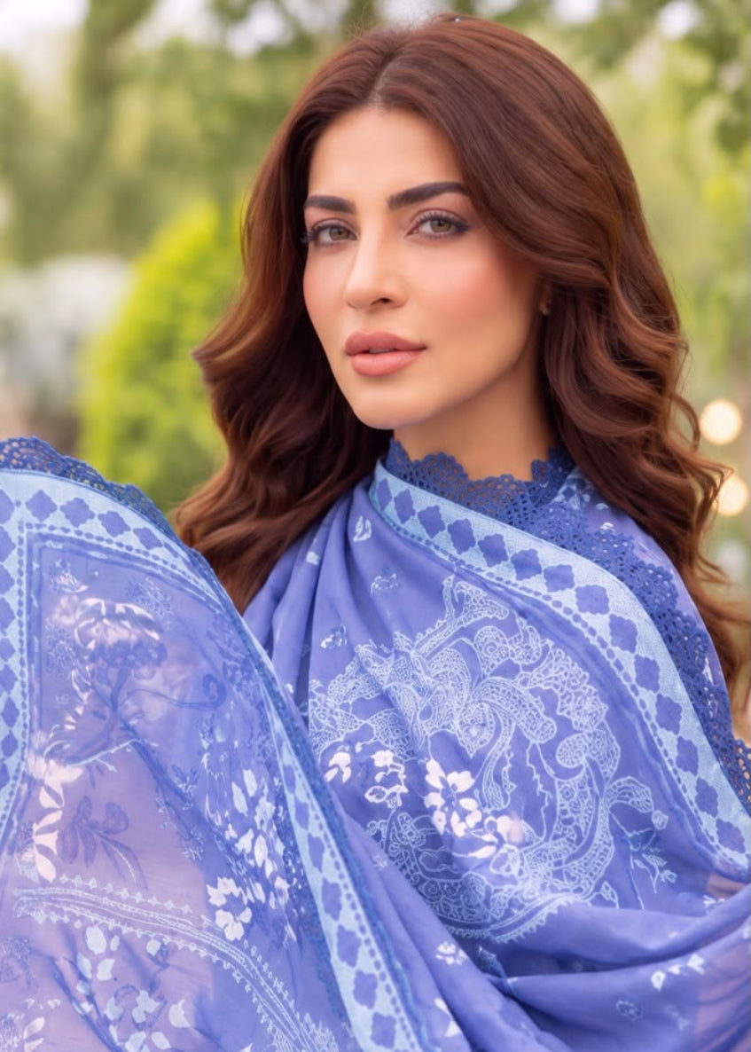 A woman wearing a sheer lavender-blue embroidered dupatta or scarf with white floral and geometric patterns, photographed outdoors against a softly blurred green background.