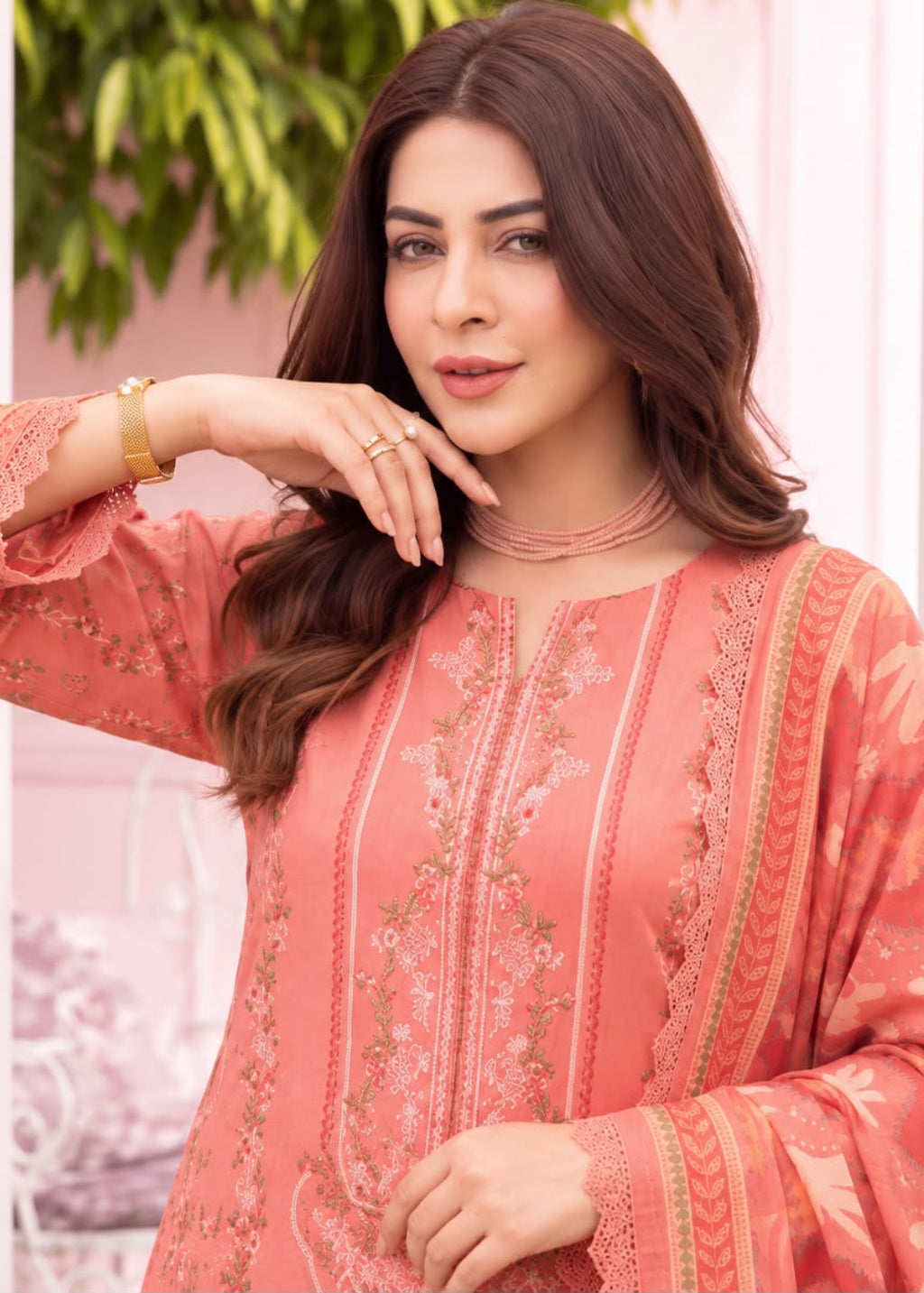 A woman wearing a peach-colored embroidered traditional outfit with long sleeves and lace detailing, posed in front of a soft pink and white decorative background.