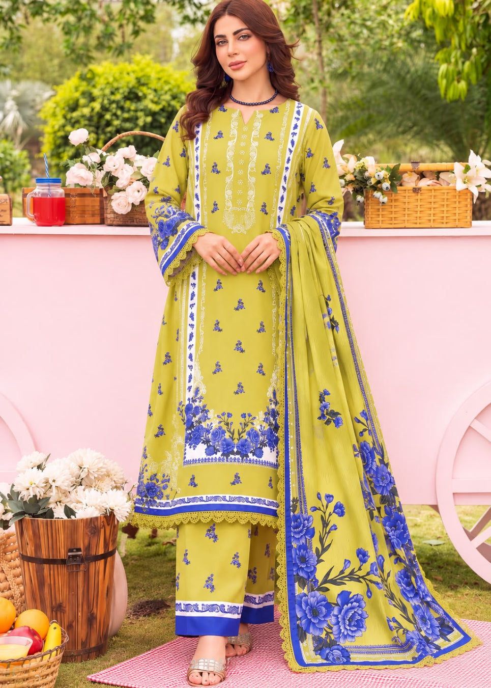 A woman models a three-piece Indian ethnic salwar kameez suit in olive green with blue floral prints and white borders. The set includes a long-sleeved tunic, matching trousers, and a coordinating dupatta, set against a soft pink and green garden backdrop.