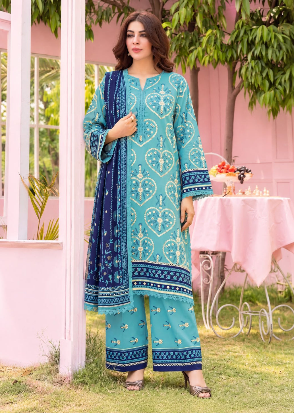 A woman models a traditional three-piece salwar kameez suit in turquoise and navy blue, featuring intricate white and blue embroidery, set against a pink house and green garden background.