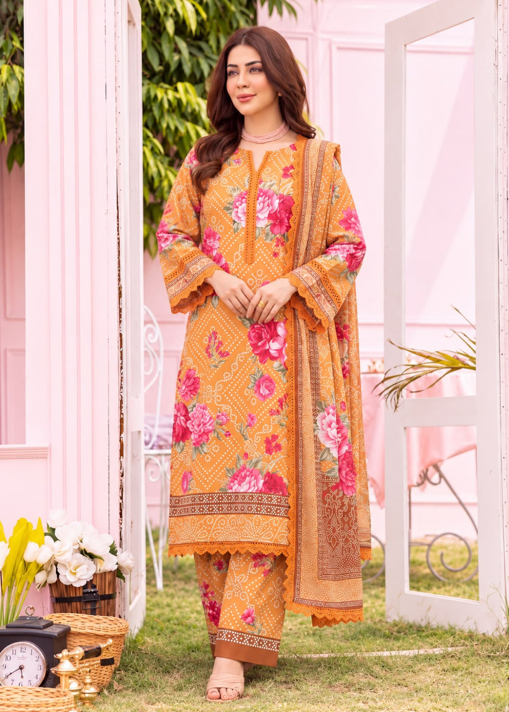 A woman models a three-piece traditional South Asian salwar kameez suit in a garden setting. The suit is mustard yellow with intricate pink and red floral prints, geometric patterns, and a matching dupatta.