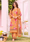 A woman models a three-piece traditional South Asian salwar kameez suit in a garden setting. The suit is mustard yellow with intricate pink and red floral prints, geometric patterns, and a matching dupatta.