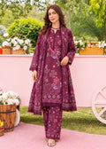 A woman models a three-piece traditional Indian salwar kameez suit in deep maroon with floral embroidery and prints, featuring a long-sleeved tunic, matching trousers, and a coordinating sheer jacket. The setting is a styled outdoor garden with a pink wall and decorative baskets.