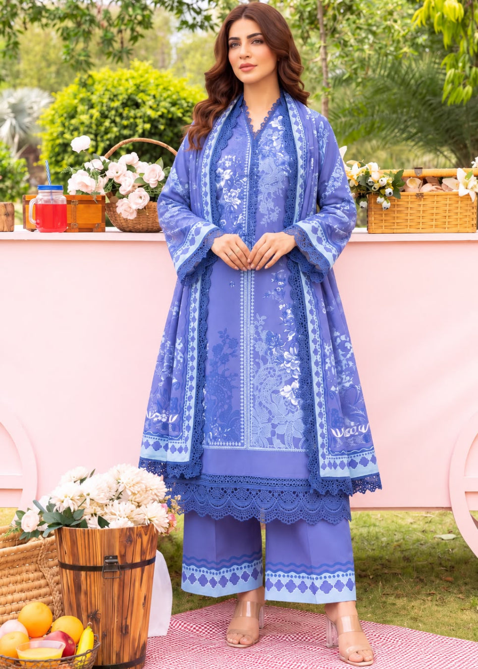 A woman models a three-piece traditional South Asian outfit in a garden setting. The outfit is a royal blue set with white floral embroidery and lace borders, consisting of a long kurta, matching trousers, and a coordinating dupatta.