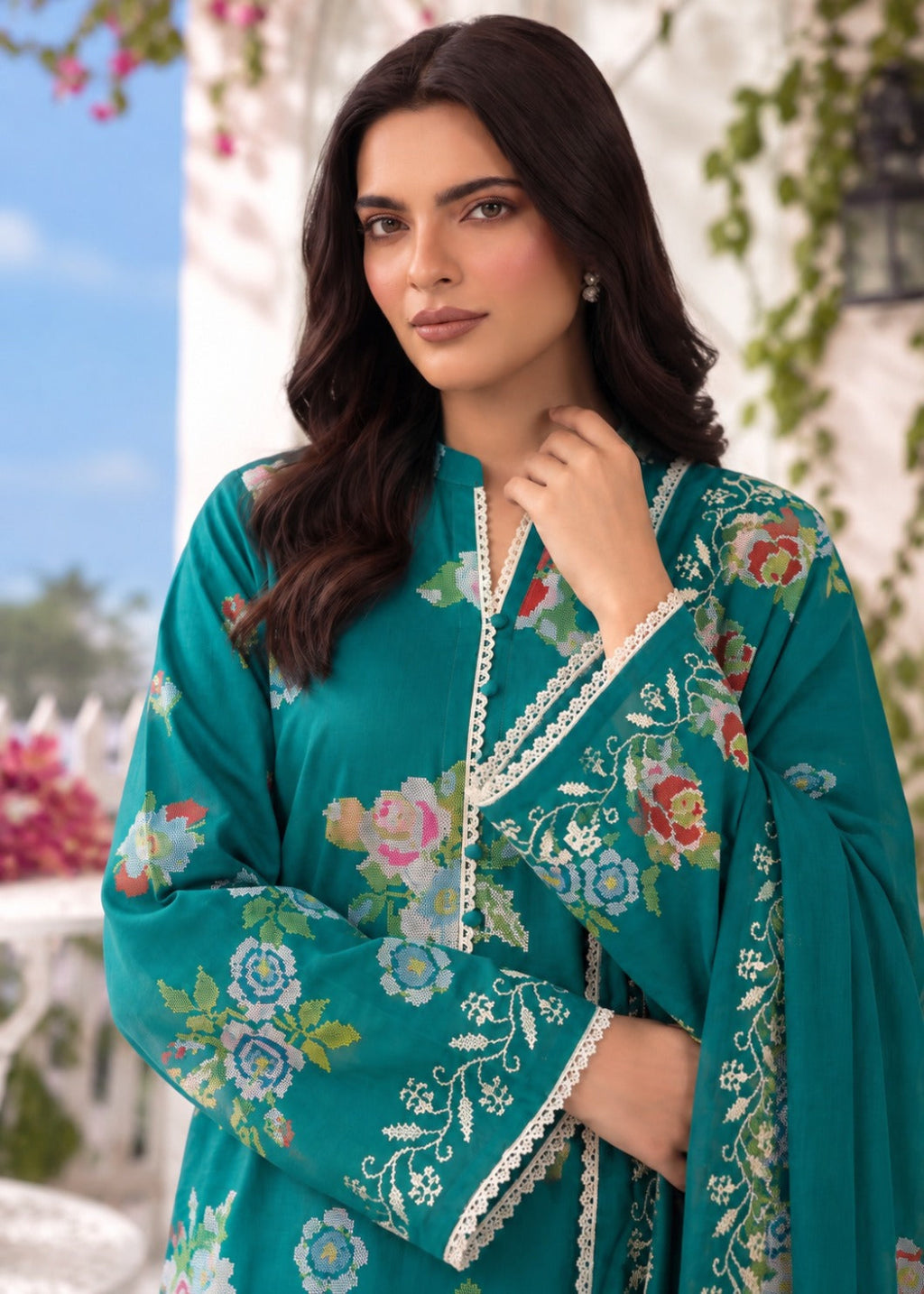A woman wearing a teal green embroidered kurta with floral patterns and white lace trim, photographed outdoors against a blurred background of white walls and greenery.
