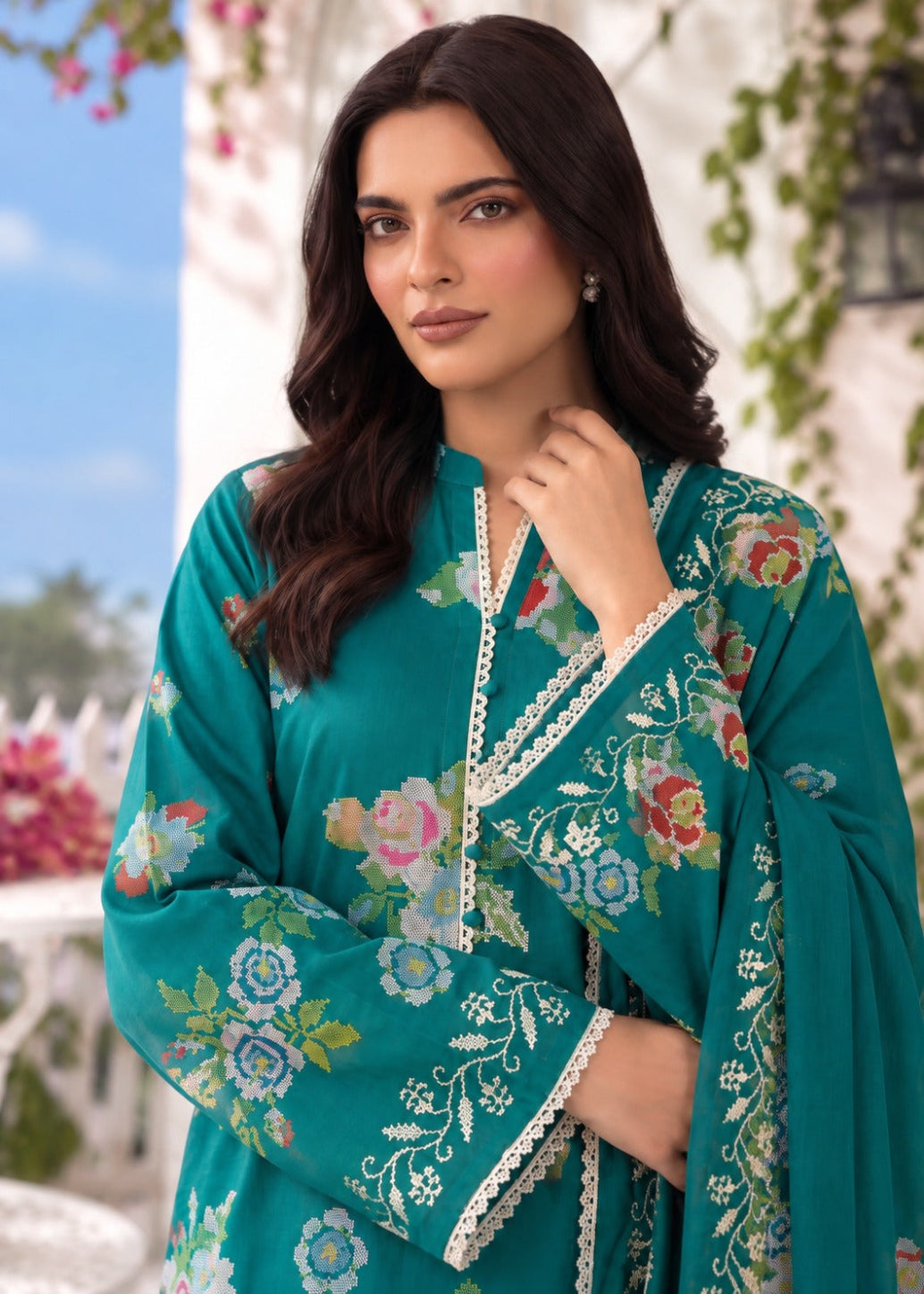 A woman wearing a teal green embroidered kurta with floral patterns and white lace trim, photographed outdoors against a blurred background of white walls and greenery.