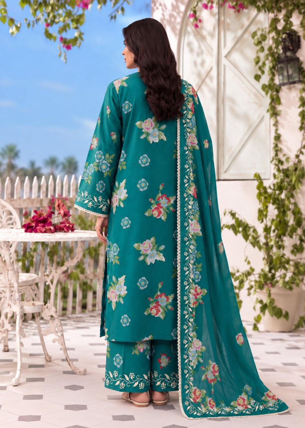 A woman stands outdoors wearing a teal floral salwar kameez suit with matching pants and a long dupatta. The outfit features pink, green, and white floral embroidery, and the background shows a patio setting with white furniture and greenery.