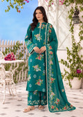 A woman stands outdoors wearing a teal floral salwar kameez suit with matching pants and a long dupatta. The outfit features pink, green, and white floral embroidery, and the background shows a patio setting with white furniture and greenery.