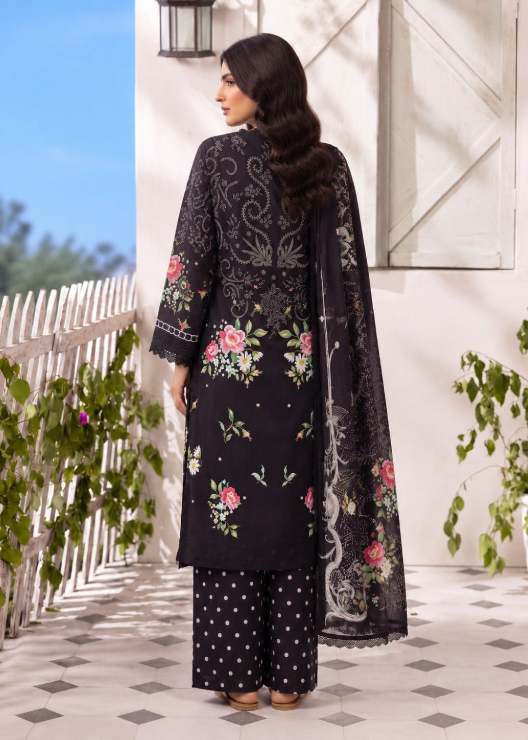 A woman modeling a black three-piece ethnic suit with floral embroidery, featuring a long kurta, matching trousers with white polka dots, and a sheer black dupatta, set against an outdoor patio backdrop.