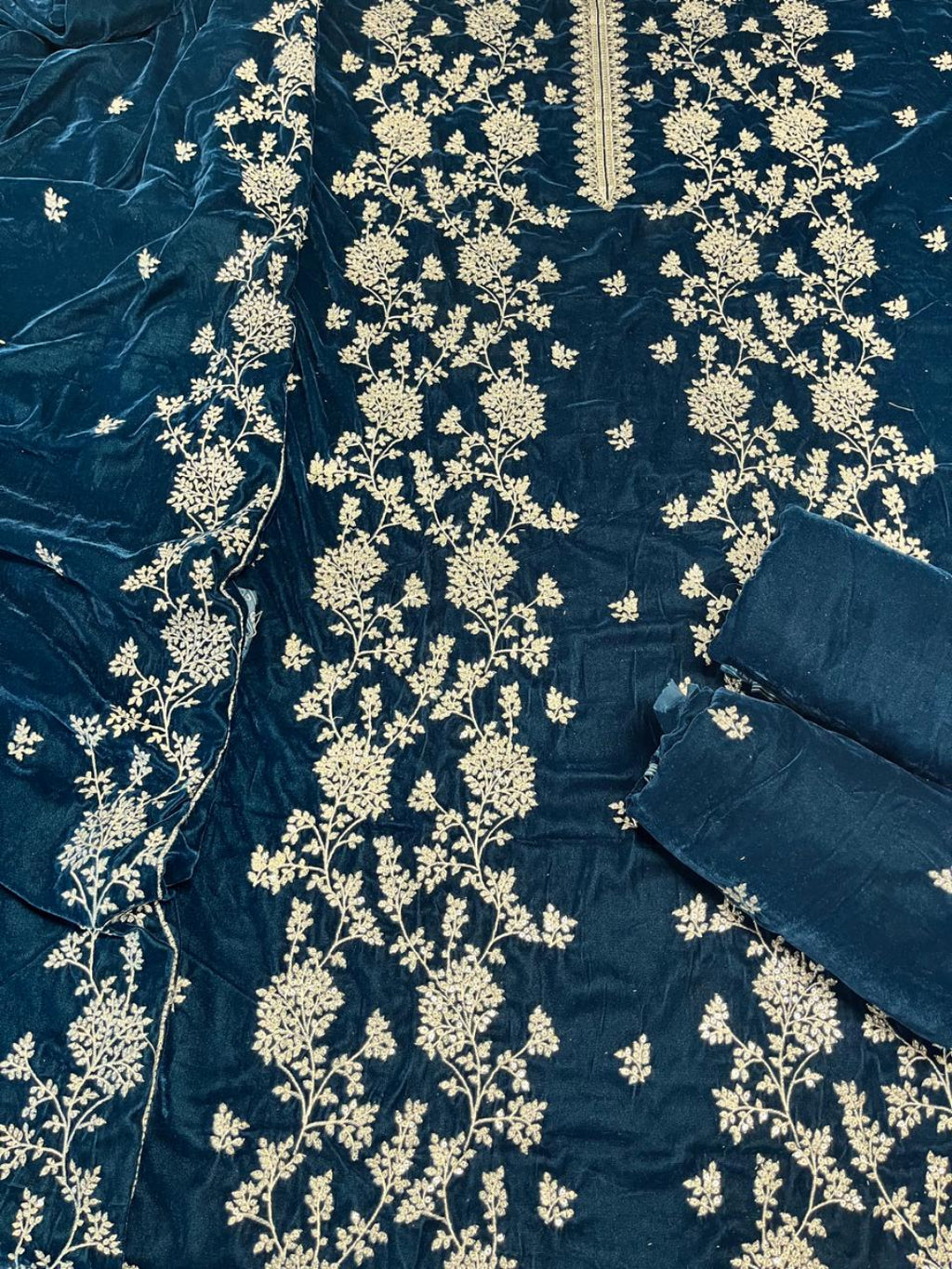 A close-up of a dark teal blue fabric garment or textile with extensive cream floral embroidery and a matching embroidered border, laid flat with a folded matching fabric piece on the right side.