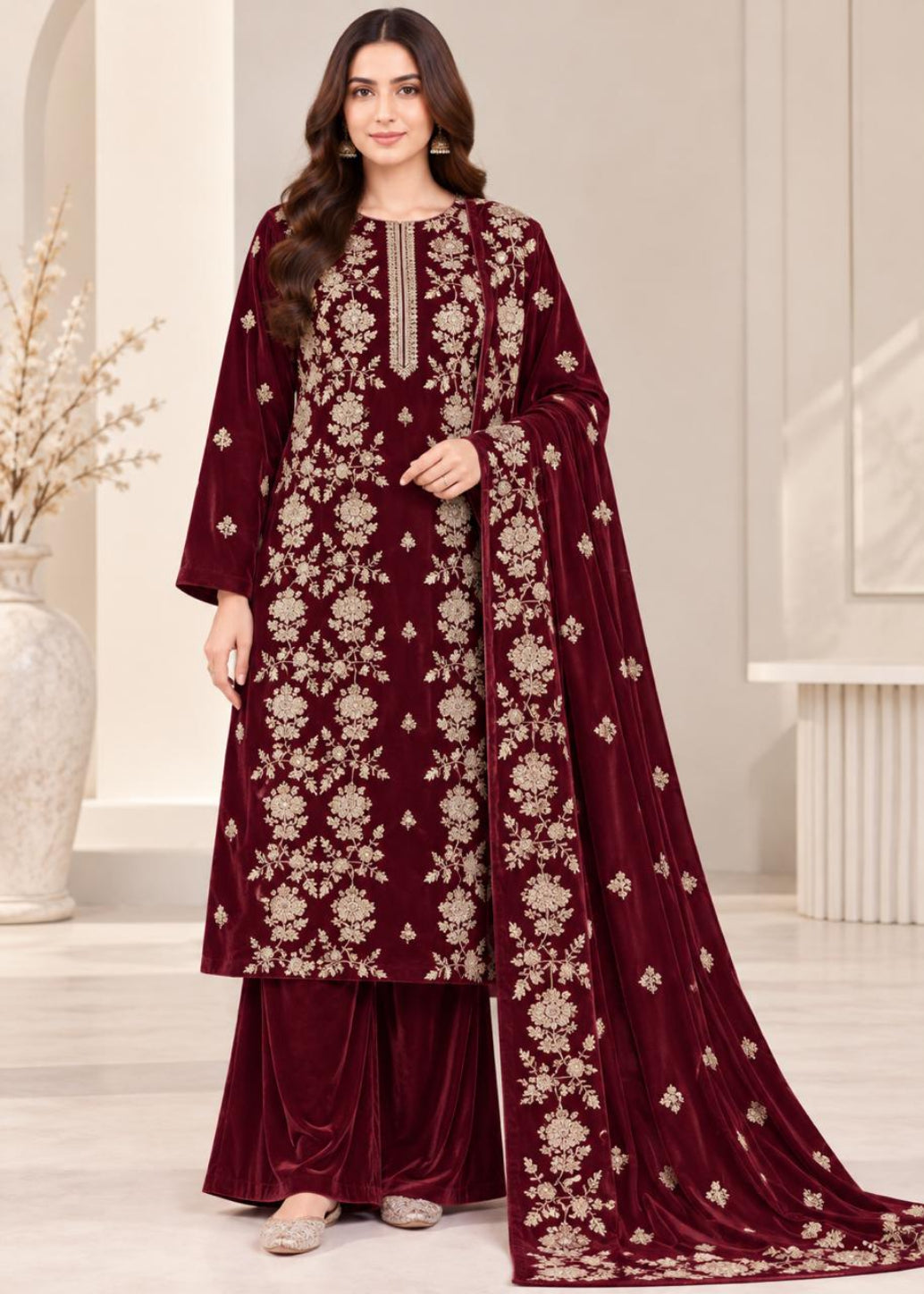 A woman models a deep maroon velvet ethnic suit with beige floral embroidery, matching pants, and a draped dupatta in a bright, modern indoor setting.