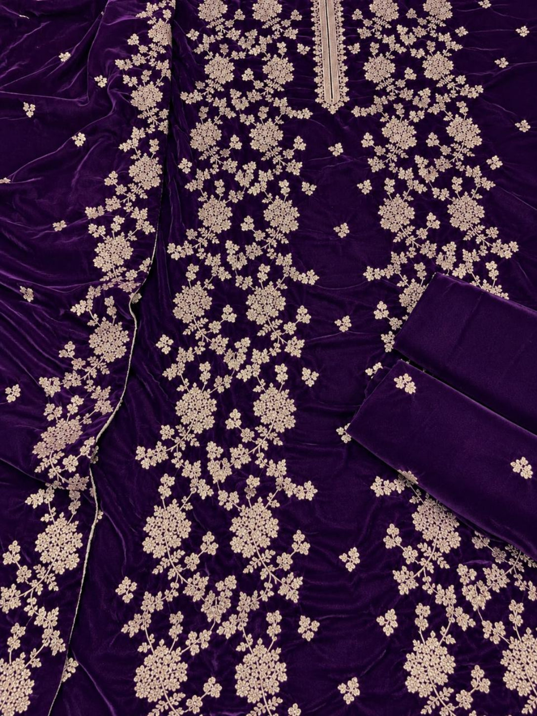 A close-up of a deep purple velvet fabric, likely a traditional ethnic garment, decorated with intricate beige floral embroidery and a matching dupatta draped over the side.