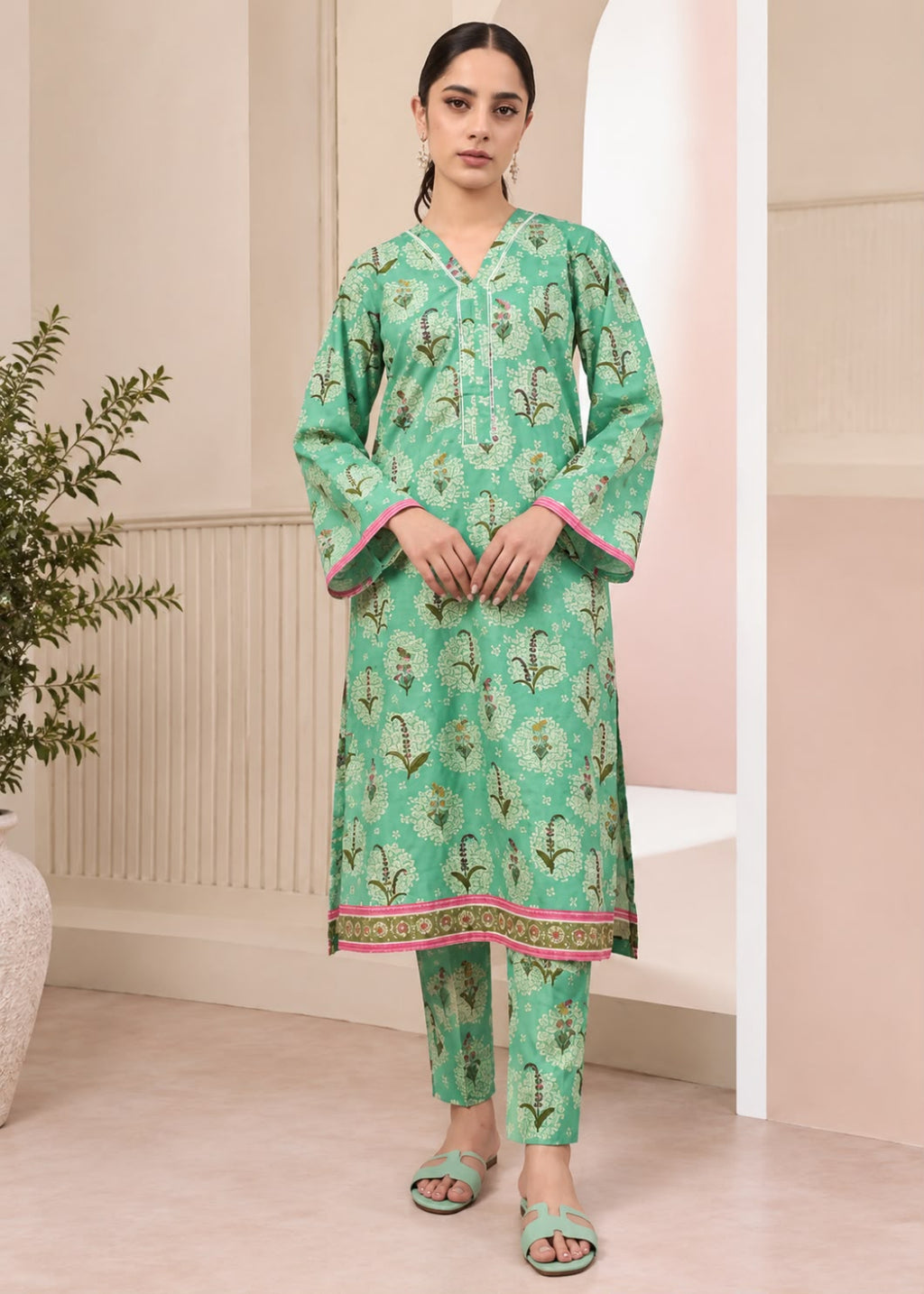 A woman modeling a mint green three-piece kurta set with floral prints and pink border accents, standing in a softly lit indoor setting with beige walls and a plant at the side.
