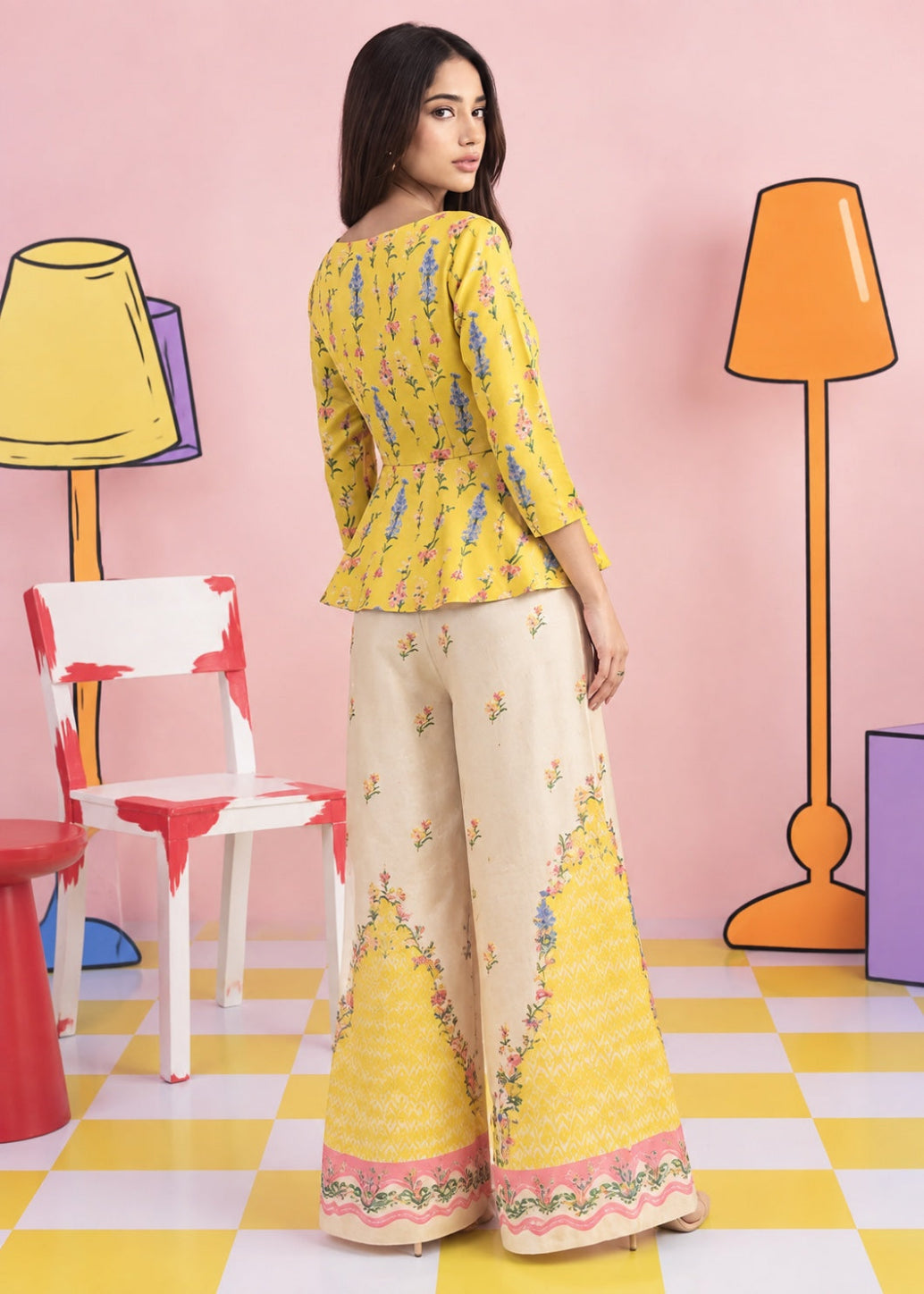 A woman modeling a yellow and beige printed traditional outfit with wide-leg pants, standing in a stylized pink room with colorful chairs and lamps.