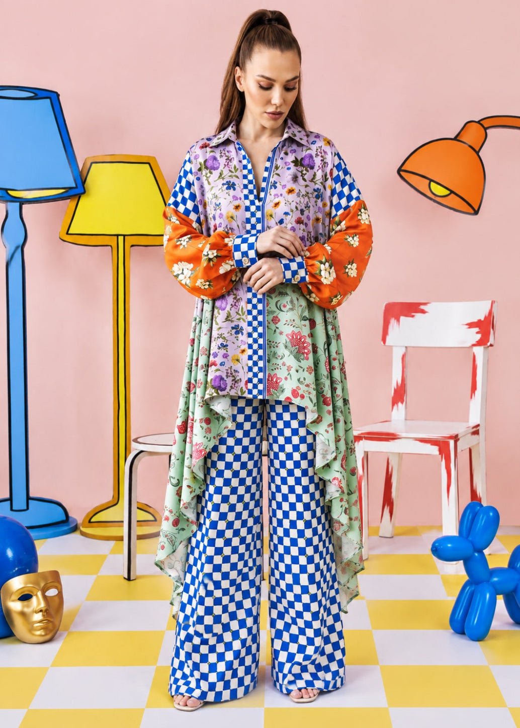 A woman stands in a stylized studio setting wearing a colorful floral longline cardigan with orange balloon sleeves and matching blue-and-white checkered wide-leg pants. The background features a pink wall, cartoon-style lamps, a chair, and a yellow-and-white checkered floor.