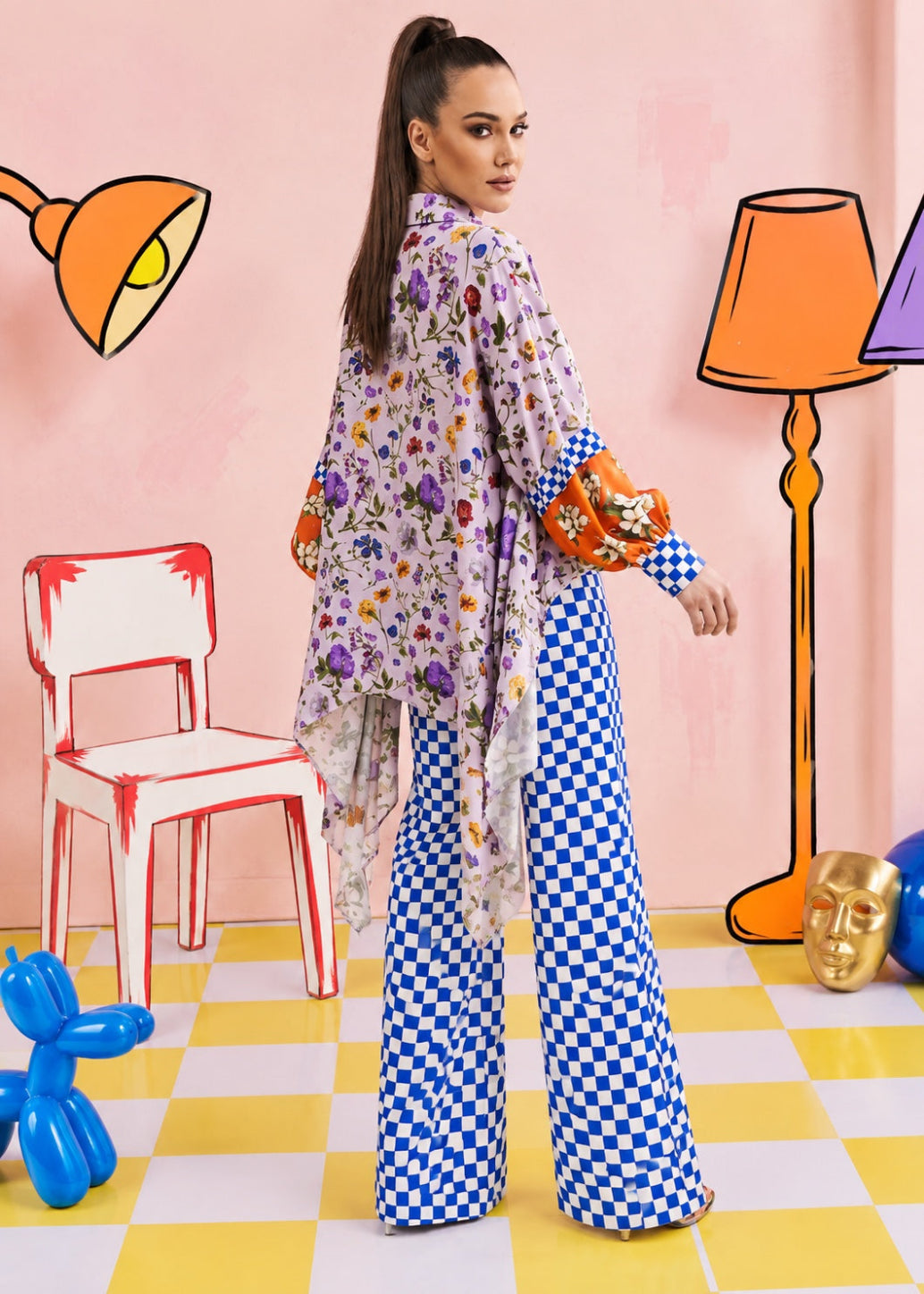 A woman stands in a stylized studio setting wearing a matching two-piece outfit: a long-sleeve button-up shirt with a multicolor floral print and wide-leg pants with a blue-and-white checkered pattern. The background features a pink wall with cartoon-style orange lamps, a red-and-white chair, a yellow-and-white checkered floor, and colorful decorative props.