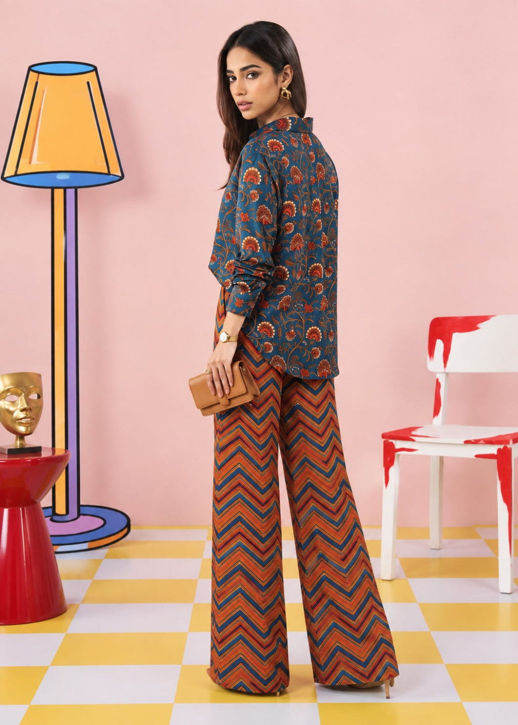 A woman modeling a two-piece printed outfit consisting of a dark blue long-sleeve top with orange and red paisley motifs and matching wide-leg trousers with a red, orange, and blue chevron pattern, set against a pink wall with colorful props.