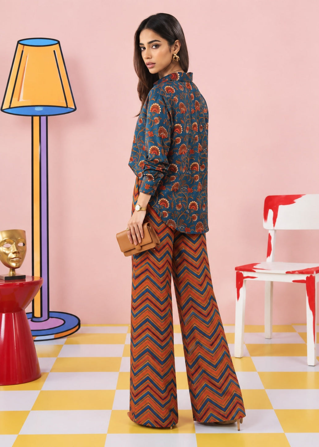 A woman modeling a two-piece printed outfit consisting of a dark blue long-sleeve top with orange and red paisley motifs and matching wide-leg trousers with a red, orange, and blue chevron pattern, set against a pink wall with colorful props.