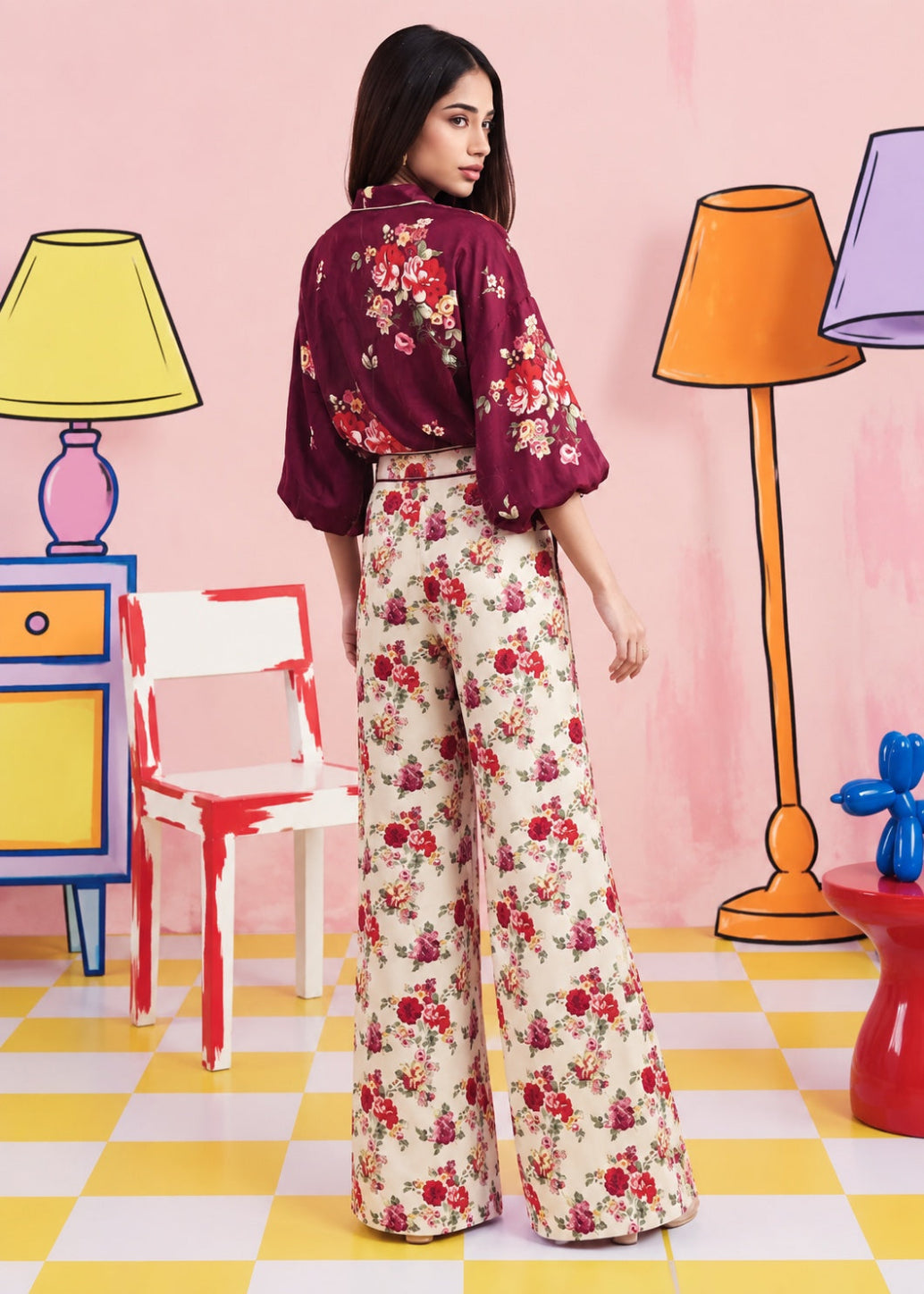 A woman stands with her back to the camera wearing a burgundy floral blouse and cream wide-leg trousers with a red floral print, set against a whimsical pink room with cartoonish lamps and furniture.