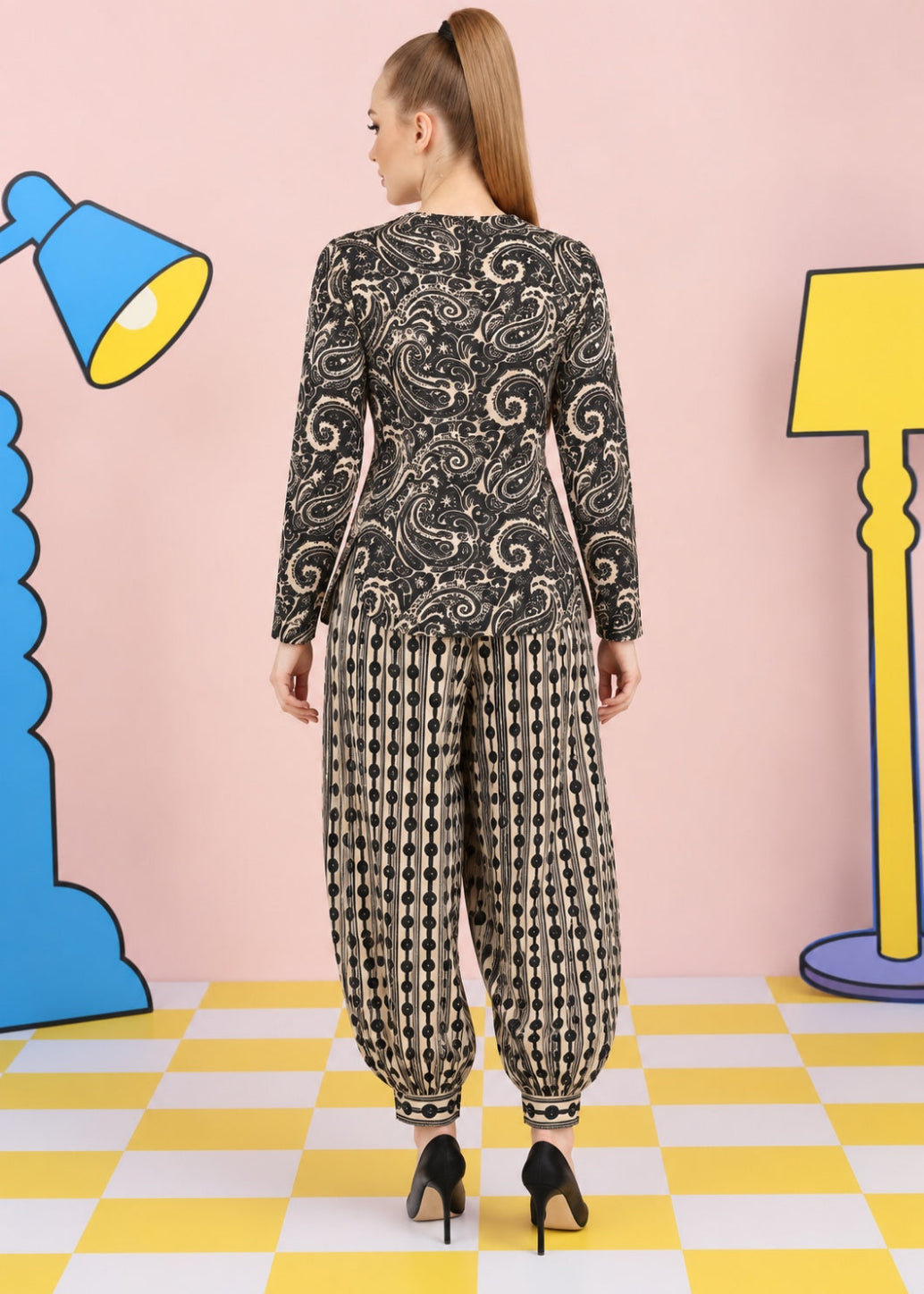 A woman stands with her back to the camera wearing a black and beige patterned long-sleeve top and matching patterned harem pants, paired with black high heels, set against a pink wall with cartoon lamp props and a yellow-and-white checkered floor.