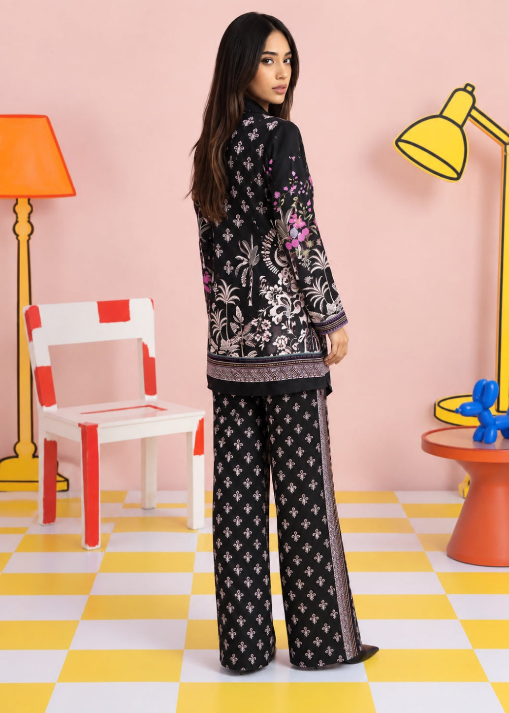 A woman stands in a stylized studio setting wearing a black two-piece outfit with white floral and geometric patterns, featuring a long-sleeve top and wide-leg pants. The background is a pastel pink wall with colorful props including a yellow lamp, a red-and-white chair, and a yellow-and-white checkered floor.