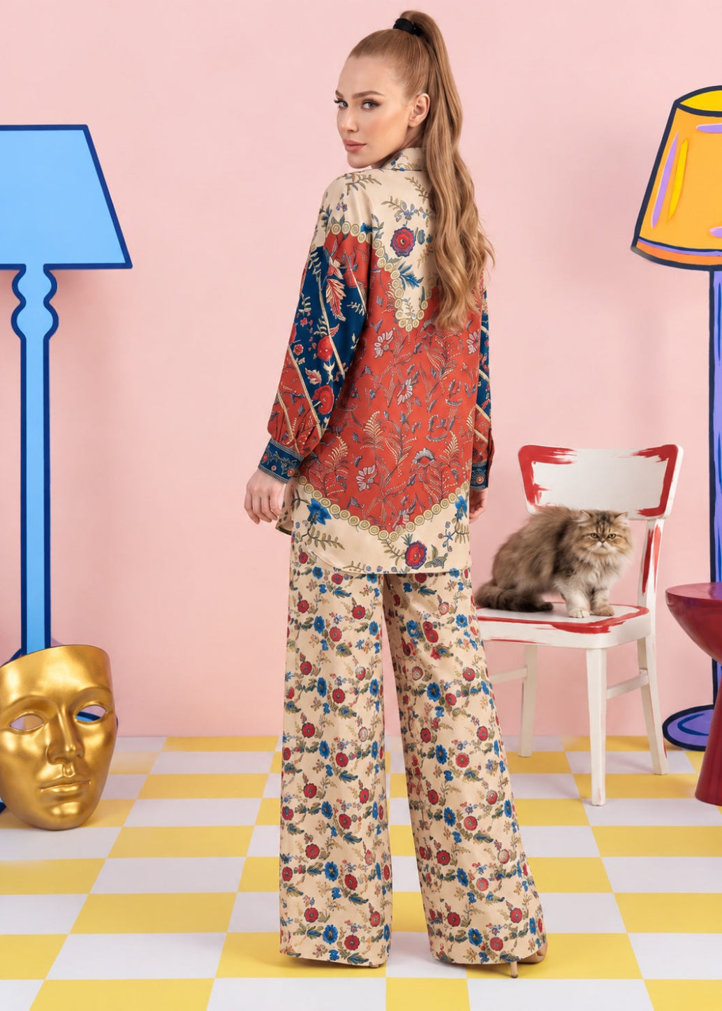 A woman stands with her back to the camera wearing a two-piece floral pajama set with a beige base and red, blue, and green patterns. The set includes a long-sleeve top and matching wide-leg pants.
