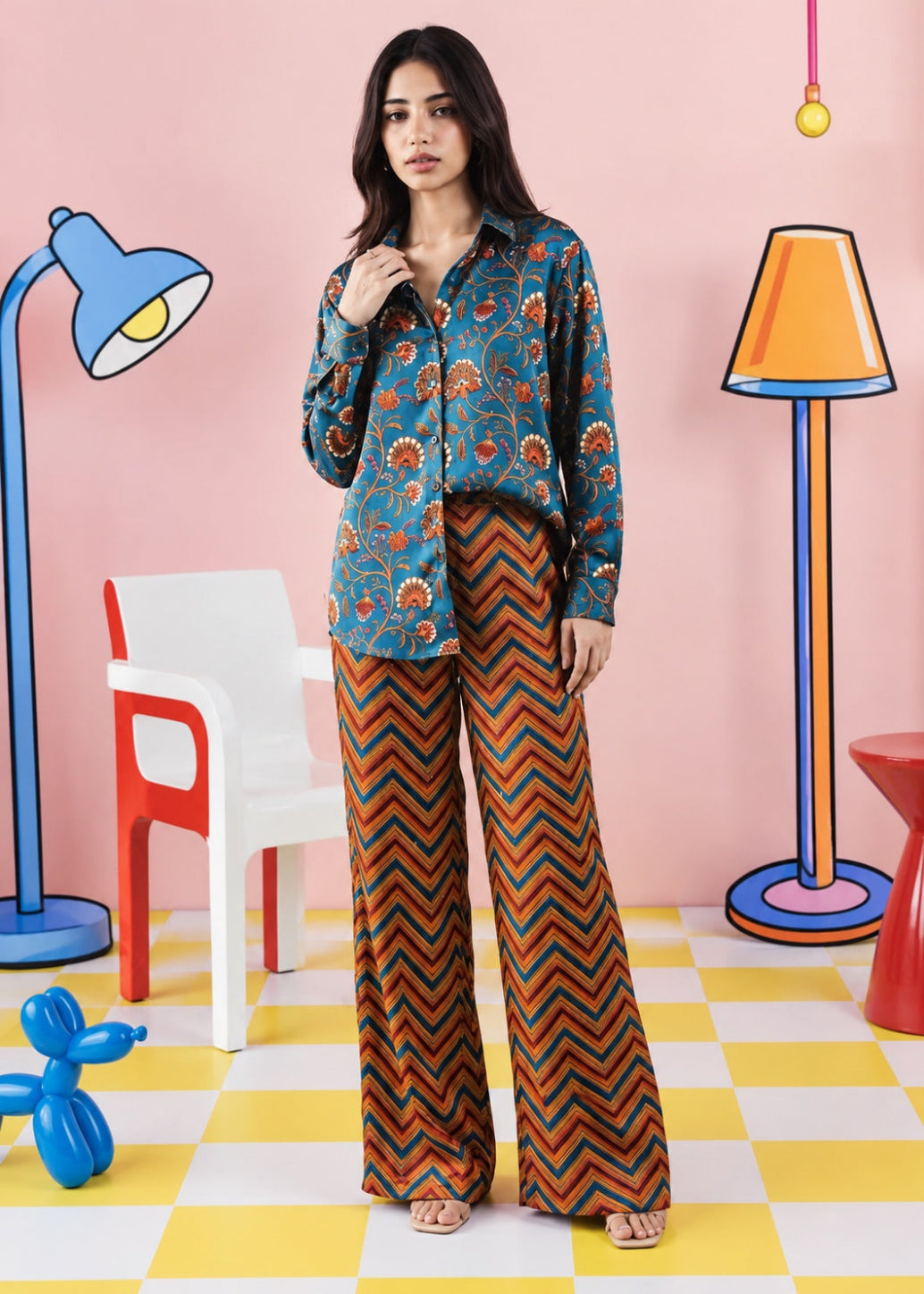 A woman stands in a stylized studio setting wearing a teal long-sleeve button-up shirt with a red and orange floral pattern, paired with wide-leg pants featuring a brown, orange, and black chevron design. The background is pink with cartoonish props including a blue floor lamp, a white chair, and a yellow-and-white checkered floor.