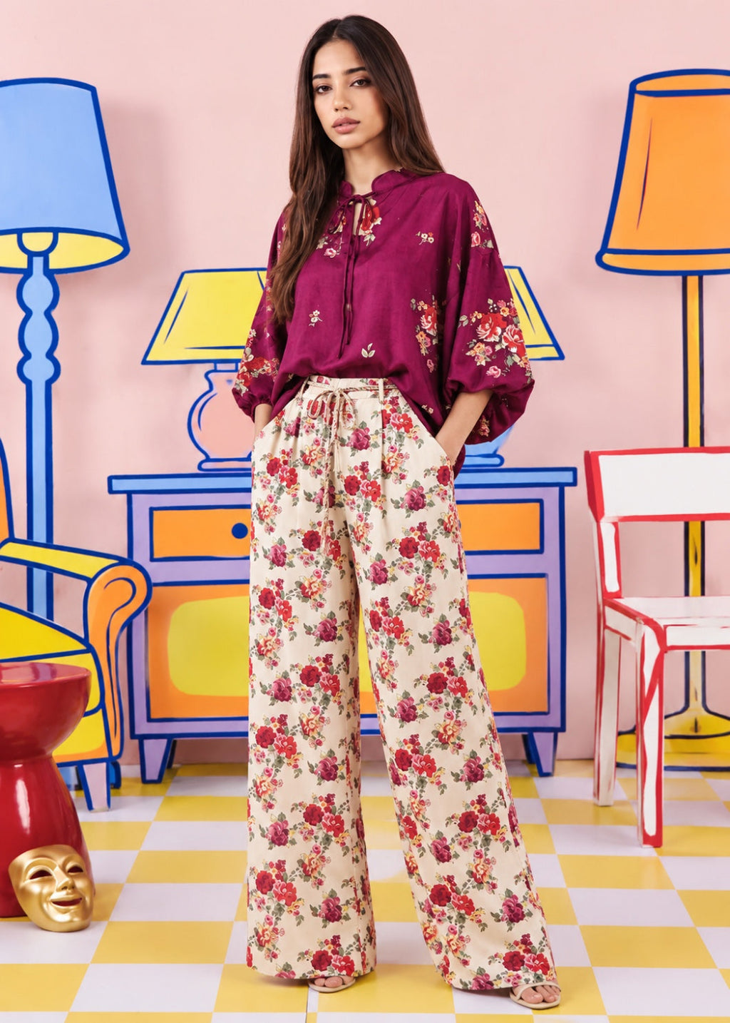 A woman stands in a stylized studio setting wearing a burgundy floral blouse and cream wide-leg trousers with a red and pink floral print. The background features colorful cartoon-style furniture and lamps on a pink wall.