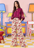 A woman stands in a stylized studio setting wearing a burgundy floral blouse and cream wide-leg trousers with a red and pink floral print. The background features colorful cartoon-style furniture and lamps on a pink wall.