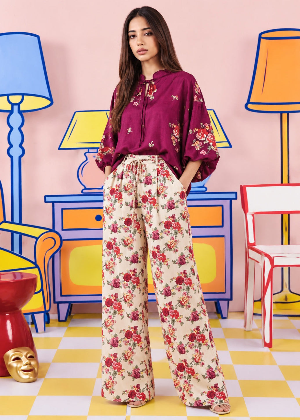A woman stands in a stylized studio setting wearing a burgundy floral blouse and cream wide-leg trousers with a red and pink floral print. The background features colorful cartoon-style furniture and lamps on a pink wall.