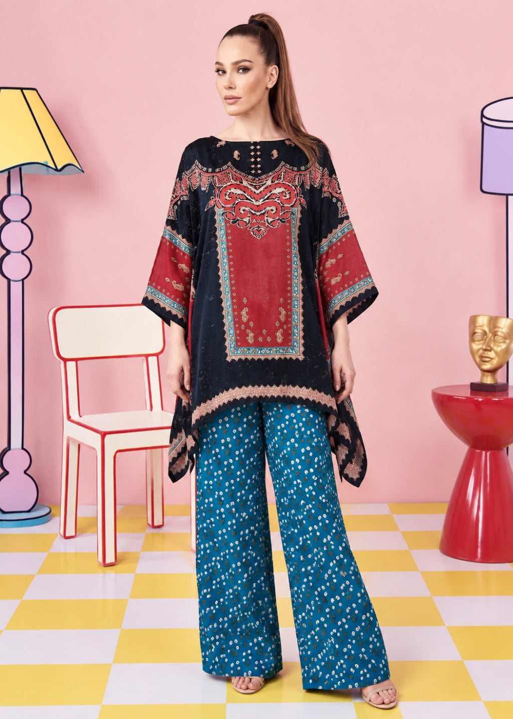 A woman models a two-piece traditional outfit featuring a black and red embroidered kurta with teal wide-leg pants, set against a stylized pink background with colorful chairs and lamps.