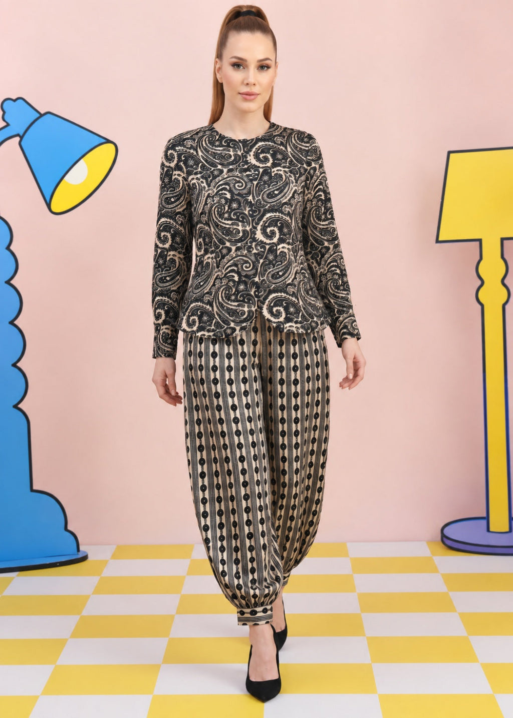 A woman modeling a black and beige paisley printed long-sleeve top with matching patterned harem pants, standing against a pink backdrop with cartoon lamp props.