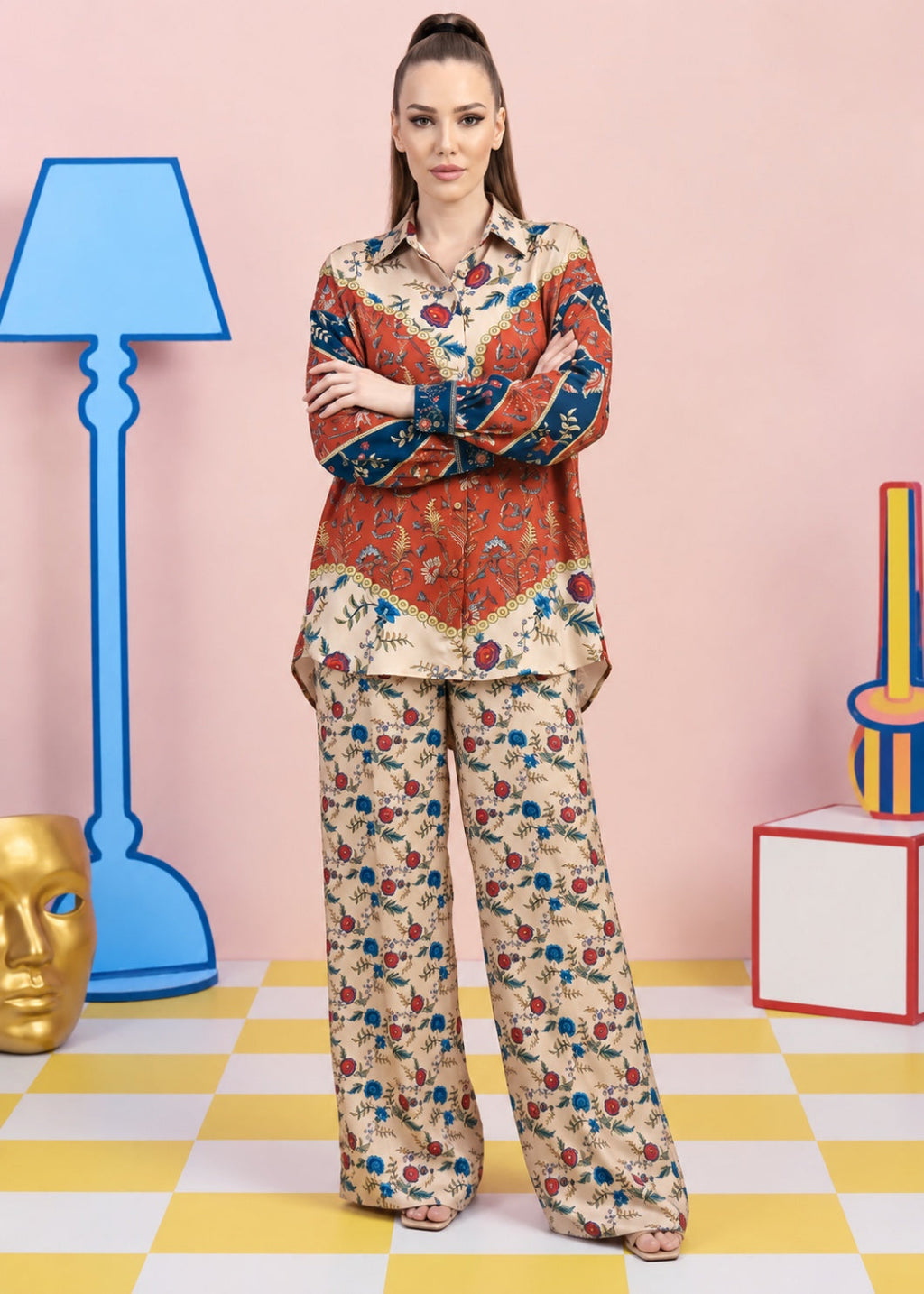 A woman stands in a studio setting wearing a matching two-piece pajama set with a beige base and a colorful floral print. The set includes a long-sleeve button-up shirt and wide-leg pants.